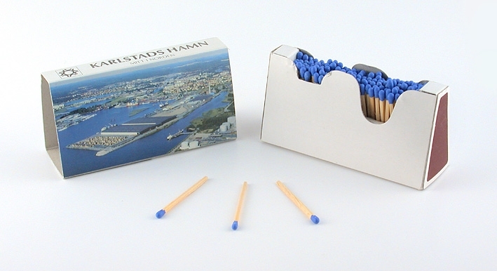 Tändsticksask av trekantig form, samt fotografier i färg på tre sidor föreställande Karlstad hamn. Stickornas huvud som ska antändas är färgade blå.