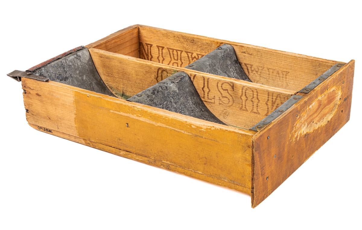 Låda av trä och plåt. Själva lådan är ihopspikad av tunna brädor vilka synes ha sågats ur en större låda för frakt el. dyl. Ljusa brädor. Framsidan av lådan är något bredare än resten: 225mm. Lådan är genom brädväggar indelad i fyra fack. De två främre något större än de bakre. Tre av facken (ej det höger bak) är klätt i plåtband, lika breda som facken, dock ej sidoväggarna i facken. Plåten följer inte fackens former utan går bara i en båge ned i facken. På brädorna inuti lådan kan man i brunt läsa "MUSTAD" och "MA GARIN"- troligen ett "R" täckt av avdelning av väggarna. Även på lådans undersida står "MUSTAD" undersidan är spikad av två brädor, en smal och en bred med ca 1/2cm glapp emellan. Ca 2/4 delar av lådans ytterkanter målade i gult. Framsidan målad brun men färgen borta i en fläck.