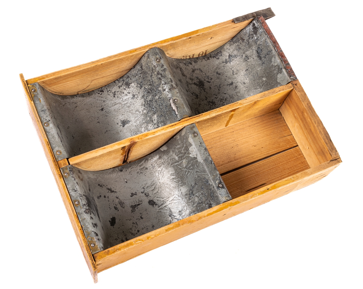 Låda av trä och plåt. Själva lådan är ihopspikad av tunna brädor vilka synes ha sågats ur en större låda för frakt el. dyl. Ljusa brädor. Framsidan av lådan är något bredare än resten: 225mm. Lådan är genom brädväggar indelad i fyra fack. De två främre något större än de bakre. Tre av facken (ej det höger bak) är klätt i plåtband, lika breda som facken, dock ej sidoväggarna i facken. Plåten följer inte fackens former utan går bara i en båge ned i facken. På brädorna inuti lådan kan man i brunt läsa "MUSTAD" och "MA GARIN"- troligen ett "R" täckt av avdelning av väggarna. Även på lådans undersida står "MUSTAD" undersidan är spikad av två brädor, en smal och en bred med ca 1/2cm glapp emellan. Ca 2/4 delar av lådans ytterkanter målade i gult. Framsidan målad brun men färgen borta i en fläck.