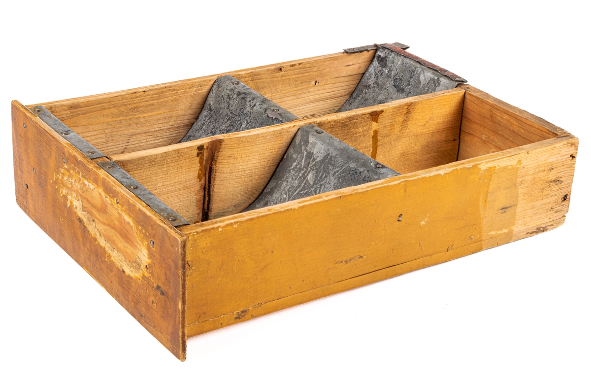 Låda av trä och plåt. Själva lådan är ihopspikad av tunna brädor vilka synes ha sågats ur en större låda för frakt el. dyl. Ljusa brädor. Framsidan av lådan är något bredare än resten: 225mm. Lådan är genom brädväggar indelad i fyra fack. De två främre något större än de bakre. Tre av facken (ej det höger bak) är klätt i plåtband, lika breda som facken, dock ej sidoväggarna i facken. Plåten följer inte fackens former utan går bara i en båge ned i facken. På brädorna inuti lådan kan man i brunt läsa "MUSTAD" och "MA GARIN"- troligen ett "R" täckt av avdelning av väggarna. Även på lådans undersida står "MUSTAD" undersidan är spikad av två brädor, en smal och en bred med ca 1/2cm glapp emellan. Ca 2/4 delar av lådans ytterkanter målade i gult. Framsidan målad brun men färgen borta i en fläck.