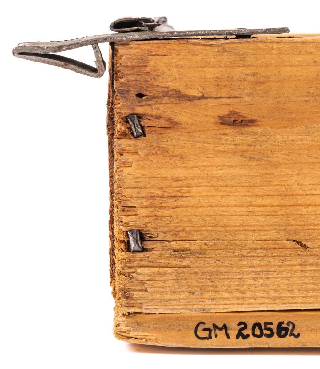 Låda av trä och plåt. Själva lådan är ihopspikad av tunna brädor vilka synes ha sågats ur en större låda för frakt el. dyl. Ljusa brädor. Framsidan av lådan är något bredare än resten: 225mm. Lådan är genom brädväggar indelad i fyra fack. De två främre något större än de bakre. Tre av facken (ej det höger bak) är klätt i plåtband, lika breda som facken, dock ej sidoväggarna i facken. Plåten följer inte fackens former utan går bara i en båge ned i facken. På brädorna inuti lådan kan man i brunt läsa "MUSTAD" och "MA GARIN"- troligen ett "R" täckt av avdelning av väggarna. Även på lådans undersida står "MUSTAD" undersidan är spikad av två brädor, en smal och en bred med ca 1/2cm glapp emellan. Ca 2/4 delar av lådans ytterkanter målade i gult. Framsidan målad brun men färgen borta i en fläck.