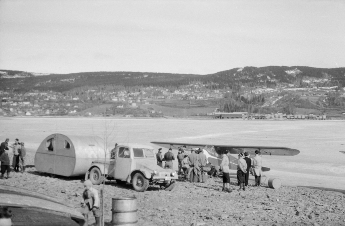 Oppland, Lillehammer, Mjøsa. Et småfly står på stranda på vestsiden av Mjøsa. Bil og campingvogn. Mange personer ser på. Utsikt mot Søre Ål med Årettadalen og Dampsaga. Bilen er en tysk Krupp feltvogn av en type som gikk under kallenavnet "maurslukeren". 