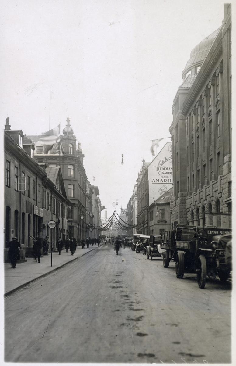 Gatebilde med fotgjengere og parkerte biler med juledekorasjoner på plass - Minnedagen for Roald Amundsen - 14. desember 1928. Fra stilheten i Oslo.