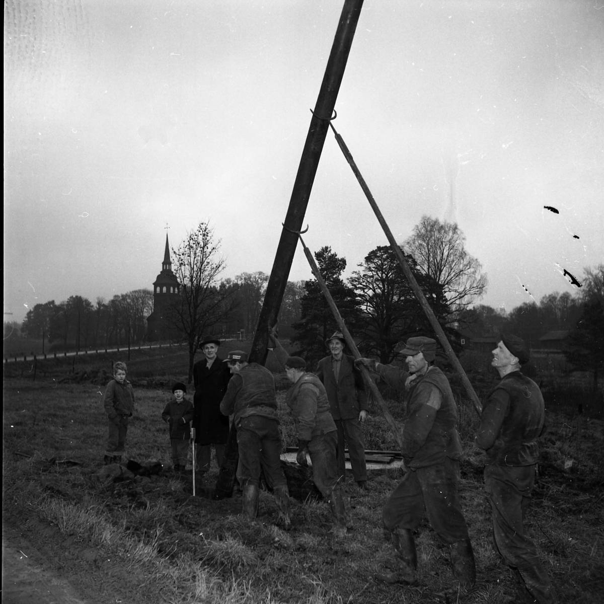 Några män arbetar med att resa en stolpe för vägbelysning i Vireda i december 1960. I bakgrunden syns Vireda kyrka.