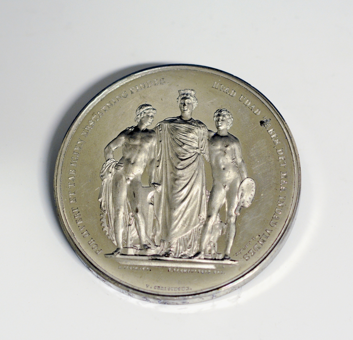 Prot: Sølvemedalje. Adversen viser en stor bygning med flagg og vimpler. Over i et bånd: "Den nordiske industri- og kunstutstilling" Under: "Kjøbenhavn 1872". Revers-motiv: En kvinde i gevandt og med krone på hodet, på hver side nakne, unge menn. Tekst: "For hvert et Tab igjen erstatning findes - Hvad utad tabes, det maa indad vindes" H. P. Holst. Under figurene "H. Øbrik. inv. 7 Schmalfeld feb. Nederst: "V. Christesen"