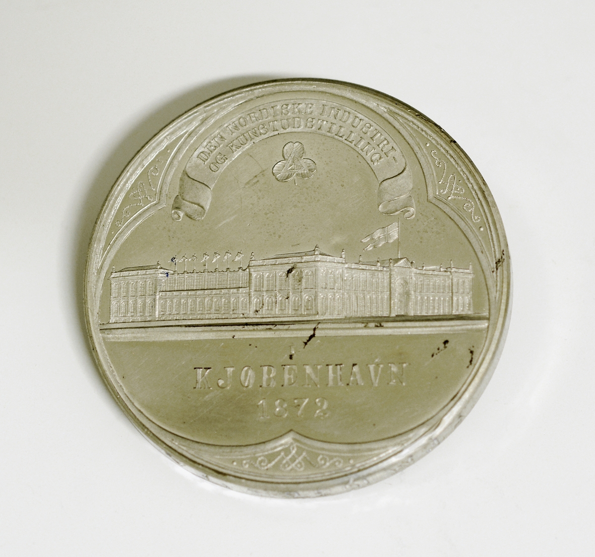 Prot: Sølvemedalje. Adversen viser en stor bygning med flagg og vimpler. Over i et bånd: "Den nordiske industri- og kunstutstilling" Under: "Kjøbenhavn 1872". Revers-motiv: En kvinde i gevandt og med krone på hodet, på hver side nakne, unge menn. Tekst: "For hvert et Tab igjen erstatning findes - Hvad utad tabes, det maa indad vindes" H. P. Holst. Under figurene "H. Øbrik. inv. 7 Schmalfeld feb. Nederst: "V. Christesen"