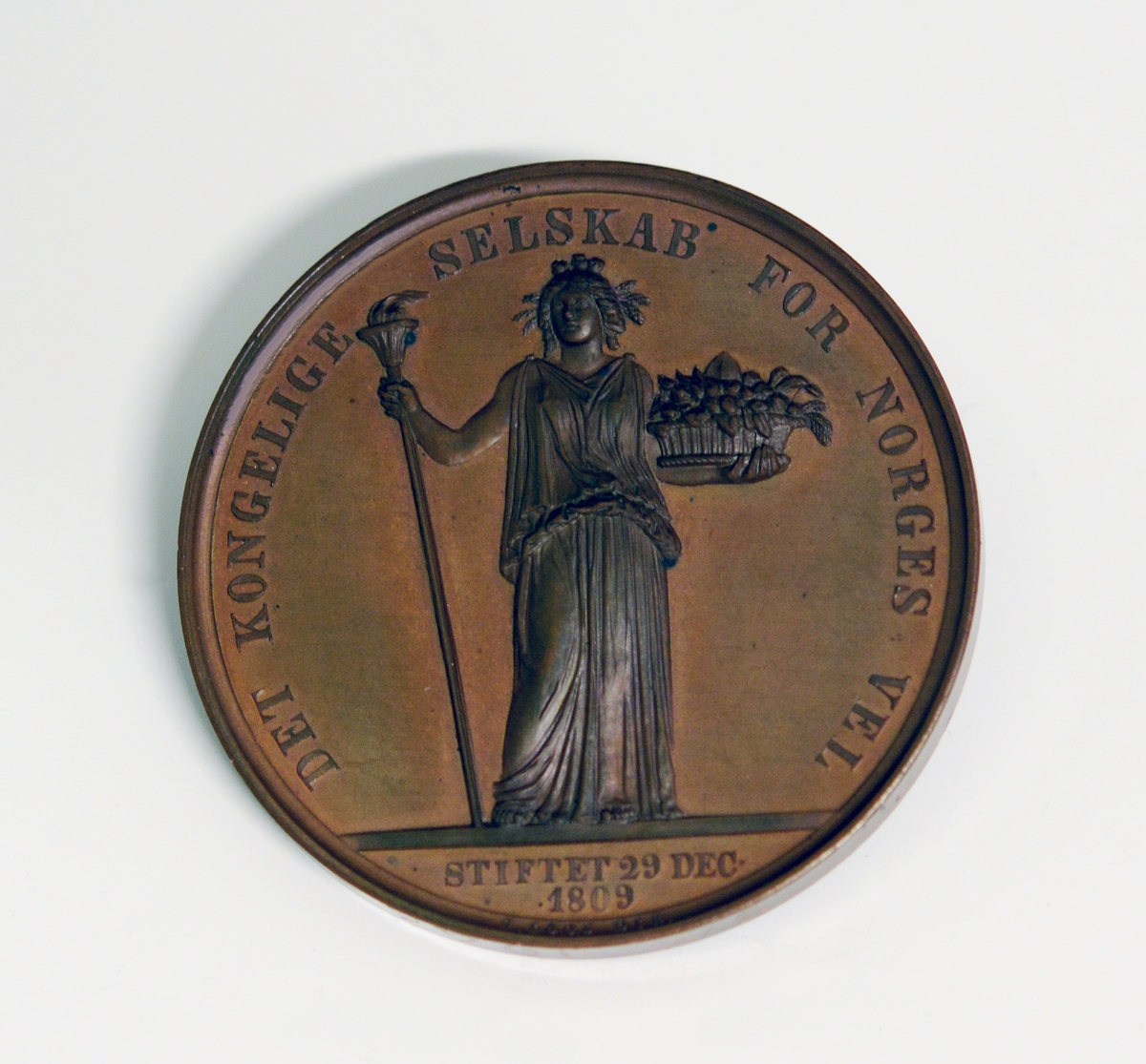 Prot: Bronsemedalje. Advers-motiv: kvinne i gevandt med fakkel i høyre hånd og en blomsterkurv i venstre hånd. Tekst: Det kongelige selskab for Norges vel Stiftet 29de dec. 1909" Under dette: "G. Loos. Dir". Revers-motiv: Laurbærkrans og over denne: "Den grønnes over grøde". På kanten inngravert "J. Jensen"