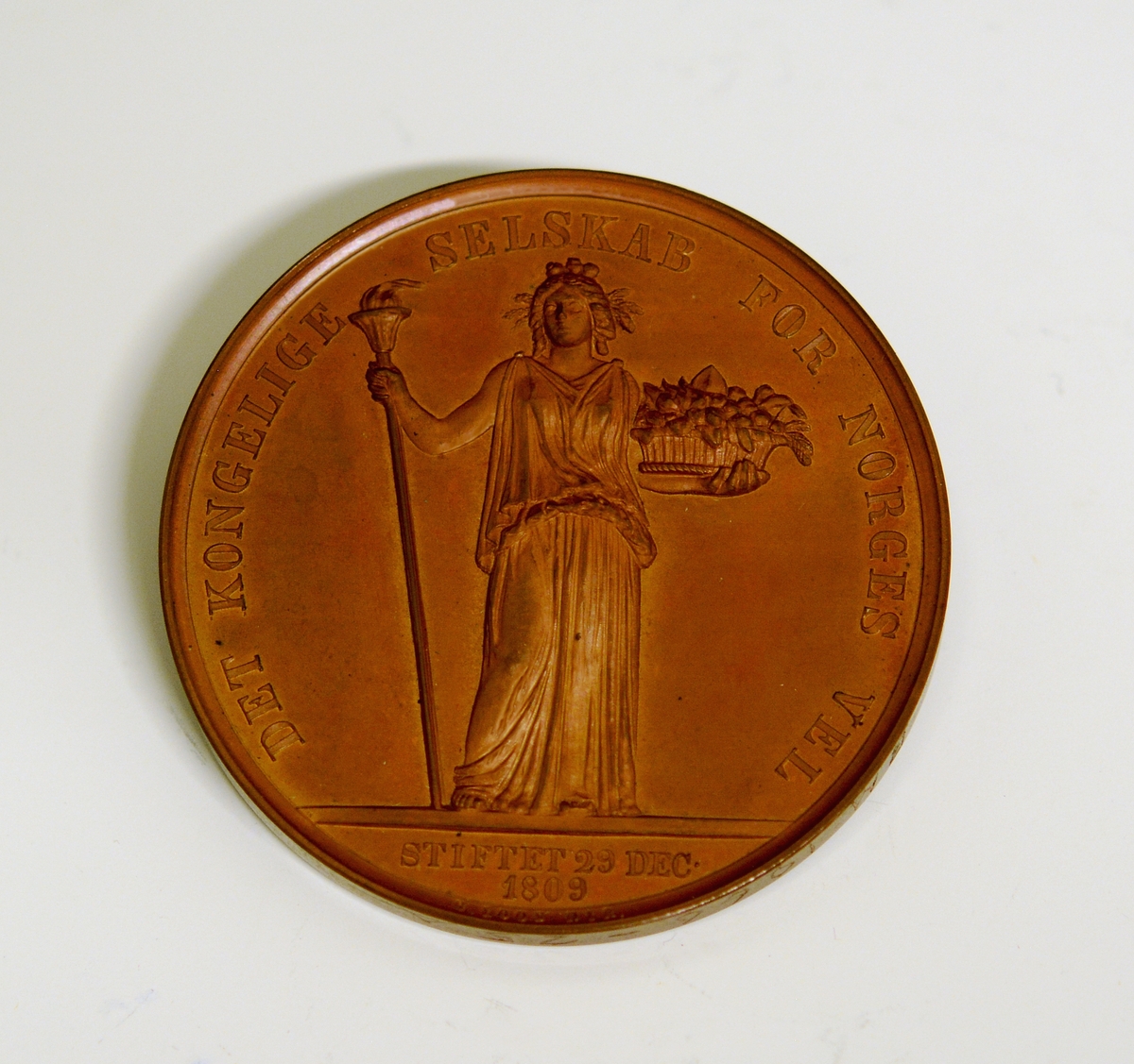 Prot: Bronsemedalje. Advers-motiv: kvinne i gevandt med fakkel i høyre hånd og en blomsterkurv i venstre hånd. Tekst: Det kongelige selskab for Norges vel Stiftet 29de dec. 1909" Under dette: "G. Loos. Dir". Revers-motiv: Laurbærkrans og over denne: "Den grønnes over grøde". På kanten inngravert "J. Jensen"
Ligger i rød firkantet eske.