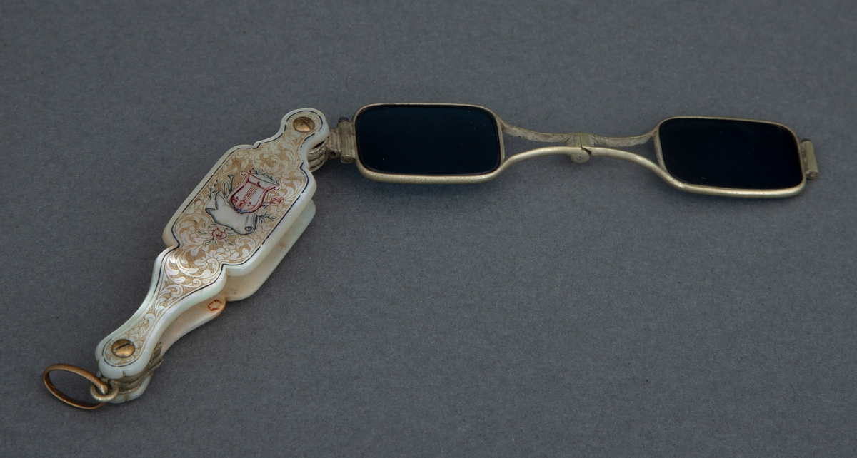 Sammenleggbar stanglorgnett i perlemorsholder. Holderens yttersider er dekorert med innrissede akantusblader på skrafert bunn og med en lyre i midten. Selve brillene er innfattet i en tynn rektangulær metallramme med avrundede hjørner. Glasset er dyptgrønt. Brillene kan ved et fjærende midtledd legges dobbelt før de skyves inn i holderen.