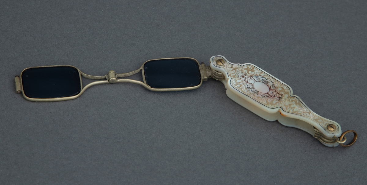 Sammenleggbar stanglorgnett i perlemorsholder. Holderens yttersider er dekorert med innrissede akantusblader på skrafert bunn og med en lyre i midten. Selve brillene er innfattet i en tynn rektangulær metallramme med avrundede hjørner. Glasset er dyptgrønt. Brillene kan ved et fjærende midtledd legges dobbelt før de skyves inn i holderen.