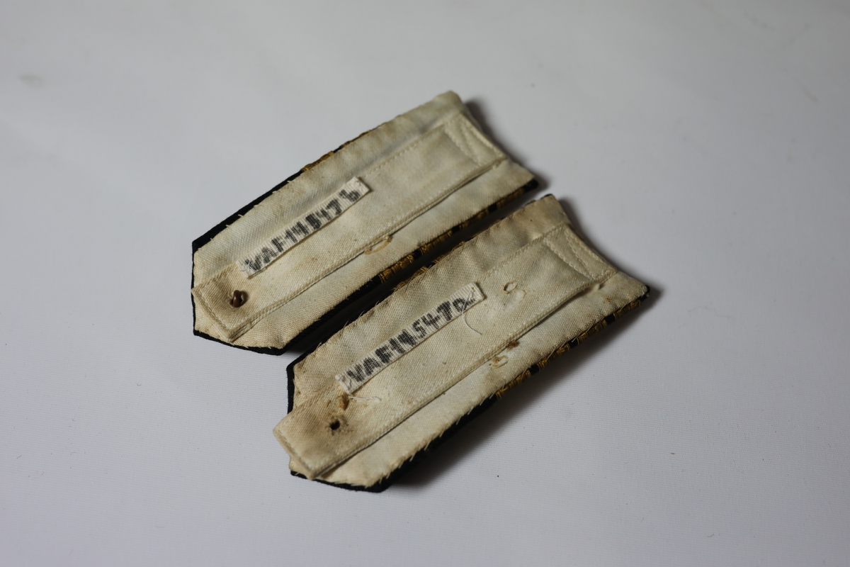 Skulderklaffer til kaptein. Den har fire gule bånd på tvers og en firkant lage av samme type bånd. I den spisse enden ved er det en gul knapp med bilde av et anker som går i gjennom en krone.