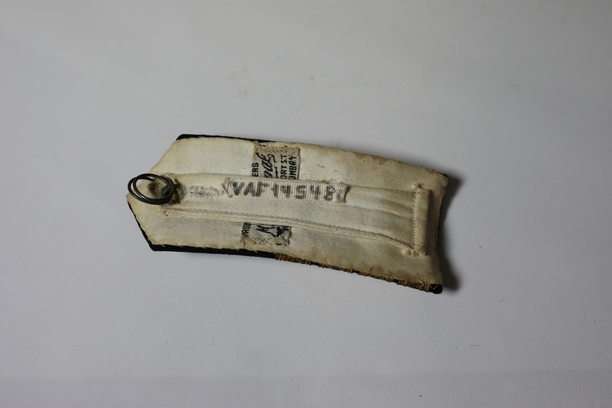 Skulderklaffer til kaptein. Den har fire gule bånd på tvers og en firkant lage av samme type bånd. I den spisse enden ved er det en gul knapp med bilde av et anker.