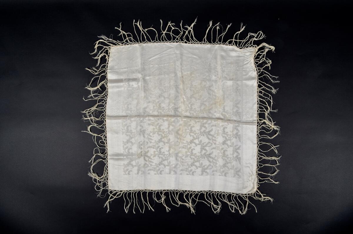 Hvitt, damaskvevd silkeskjerf med blomster- og bladmønster. Frynser på fire sider.