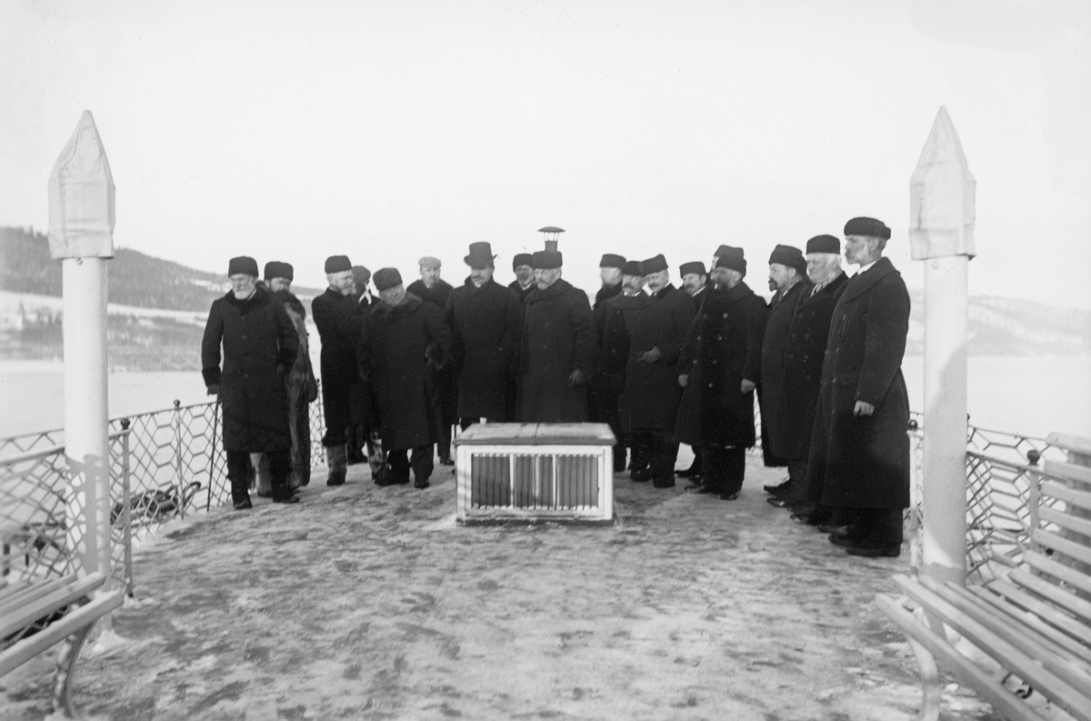 Befaring på Mjøsa i desember 1906. Fotografiet er tatt på akterdekket på en av de store dampbåtene på Mjøsa, muligens Kong Oscar, der det sto 18 menn, kledd i mørke vinterfrakker og med luer eller hatter på hodet. Bildet er fra Glomma fellesfløtingsforenings arkiv, og det er nærliggende å anta at disse mennene var rekruttert fra direksjonen (styret) og administrasjonen i Christiania Tømmerdirektion (Øvre Glommen Fellesfløtningsforening), som i 1936-37 ble fusjonert med Fredrikstad Tømmerdirektion (Nedre Glommens Fellesfløtningsforening) til Glåma Fellesfløtningsforening, som raskt ble til Glomma fellesfløtingsforening.