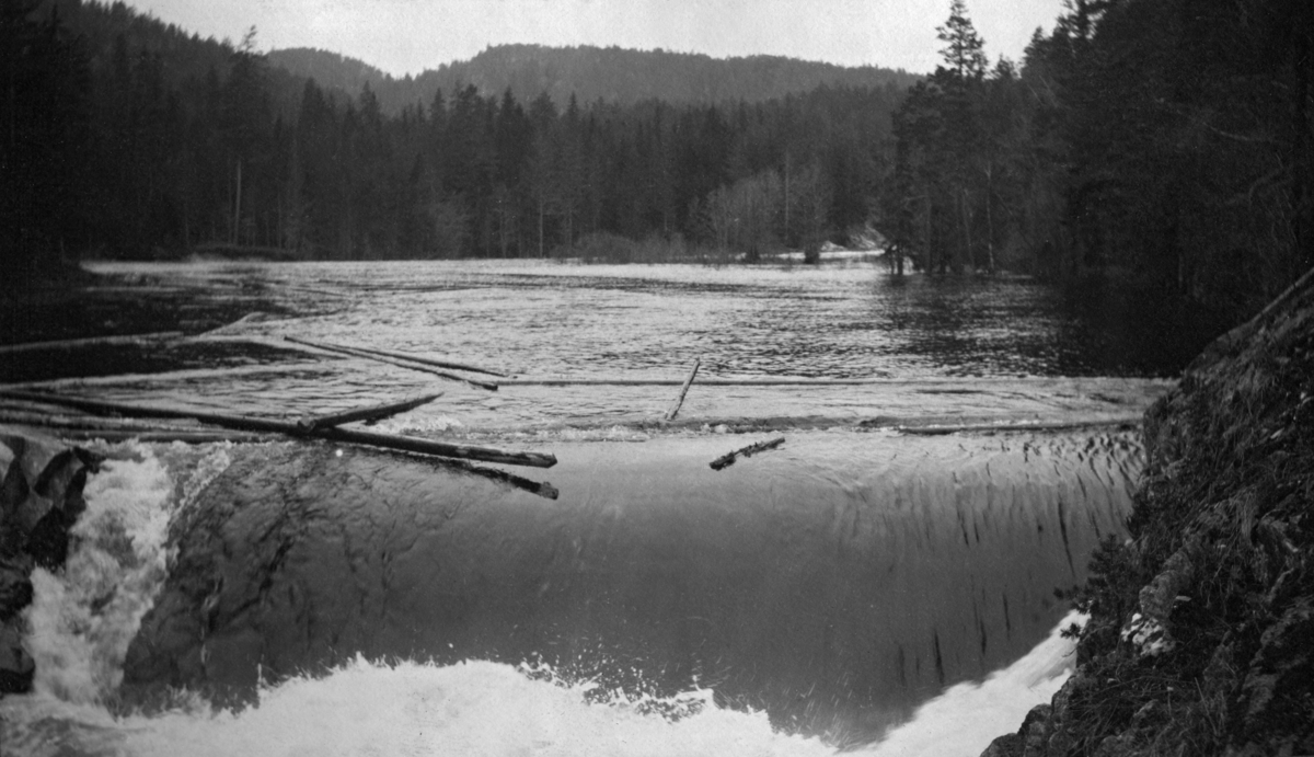 Tømmerfløting i Fulldøla, et cirka to kilometer langt elveløp fra Follsjø til Tinnåa i Notodden kommune i Telemark.  Fotografiet later til å være tatt fra ei bru mot et sted der elva – etter å ha gått i et forholdsvis bredt løp ovenfor – smalner idet den passerer en liten bergskrent i et cirka en meter høyt fossefall.  Akkurat da fotografiet ble tatt var et par tømmerstokker i ferd med å passere denne terskelen.  Fulldøla har et nedslagsfelt på 115 kvadratkilometer, og i dette området vokser det mye skog, så elva var ei viktig fløtingselv, sjøl om forholdene, som dette bildet viser, ikke var de beste.  Vanskelige fløtingsforhold ble kompensert ved regulering av Follsjå og bygging av ei 500 meter lang tømmerrenne.  Fotografiet er tatt i forbindelse med ei fløtingsbefaring sommeren 1921. 