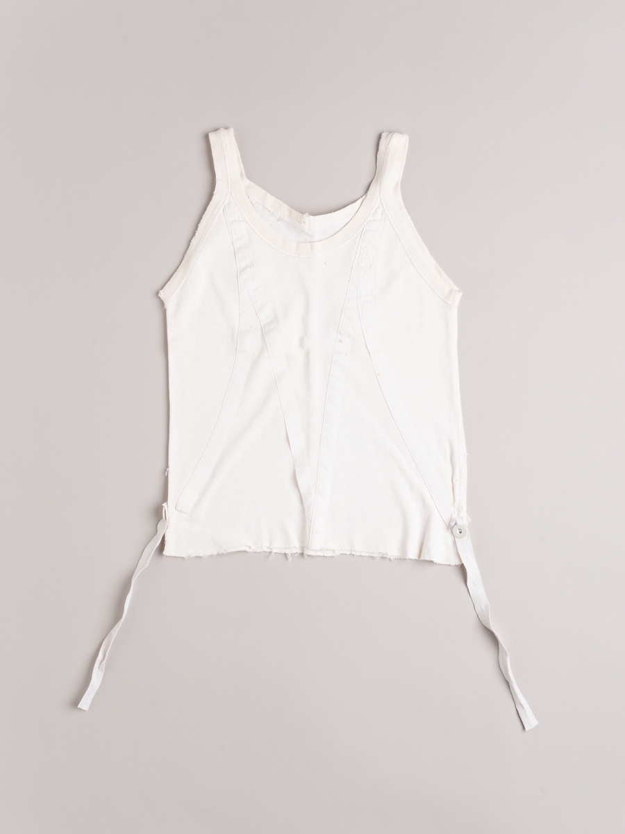 Liv (underliv) i hvit trikot. Forsterket med bendelbånd på skrå. En knapp i hver side til feste for strømpestrikk. Smale skulderstropper.
Baby- og barnetøy som først var brukt av tre søstre i USA på 1930- og 40-tallet, før det ble sendt til Norge og brukt av tre søstre i Eidsberg på 1940- og 50-tallet - kusiner av de tre i USA.