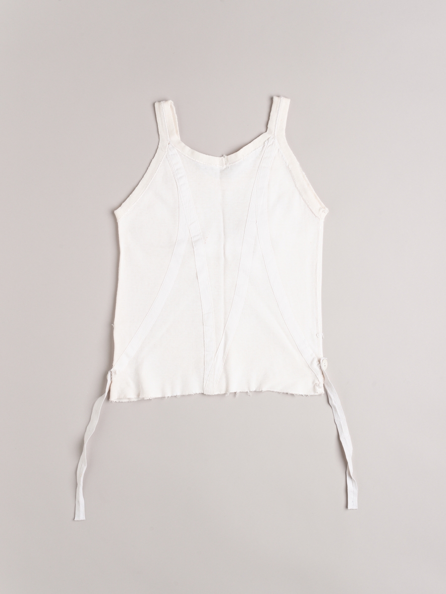 Liv (underliv) i hvit trikot. Forsterket med bendelbånd på skrå. En knapp i hver side til feste for strømpestrikk. Smale skulderstropper.
Baby- og barnetøy som først var brukt av tre søstre i USA på 1930- og 40-tallet, før det ble sendt til Norge og brukt av tre søstre i Eidsberg på 1940- og 50-tallet - kusiner av de tre i USA.