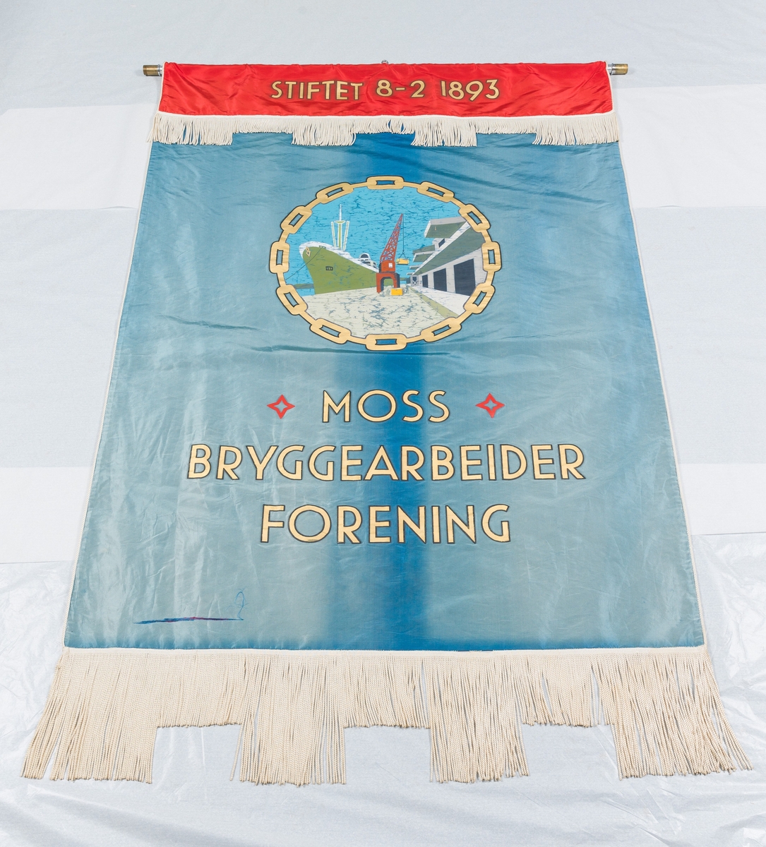 Blå fane med midtstilt motiv av skip ved kai. Rød fanekappe. Faneduken er montert på tverrstang med toppsyd og sidespyd. Bærestang.