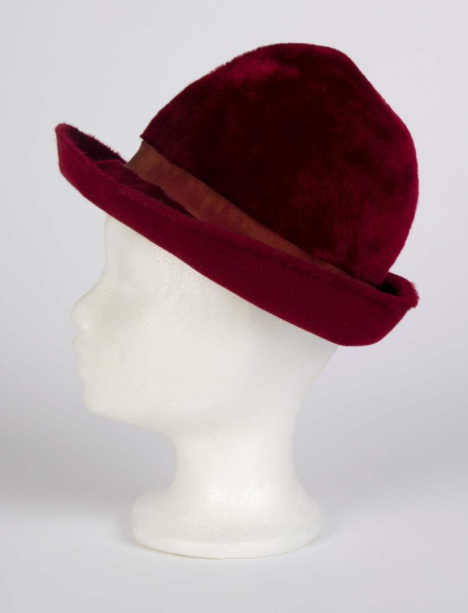 Håndsydd, burgunderrød fedora med høy, rund pull, nestet på innsiden, slik at den får to bulker på siden foran à la fedora herrehatter. Også toppen av den høye, runde pullen, er nestet på innsiden slik at den trekkes ned i en bulk. Relativt lang silkefloss. Smalt ripsbånd omkring pullen, med dekorativt element der båndet møtes bak på høyre side. Relativt bred brem, mykt oppoverbøyd og med smal falding på undersiden. Innvendig svettebånd av ripsbånd.