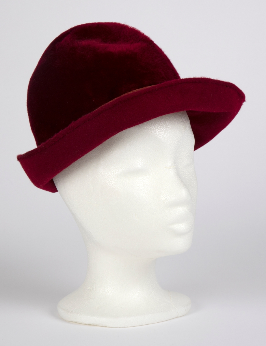 Håndsydd, burgunderrød fedora med høy, rund pull, nestet på innsiden, slik at den får to bulker på siden foran à la fedora herrehatter. Også toppen av den høye, runde pullen, er nestet på innsiden slik at den trekkes ned i en bulk. Relativt lang silkefloss. Smalt ripsbånd omkring pullen, med dekorativt element der båndet møtes bak på høyre side. Relativt bred brem, mykt oppoverbøyd og med smal falding på undersiden. Innvendig svettebånd av ripsbånd.