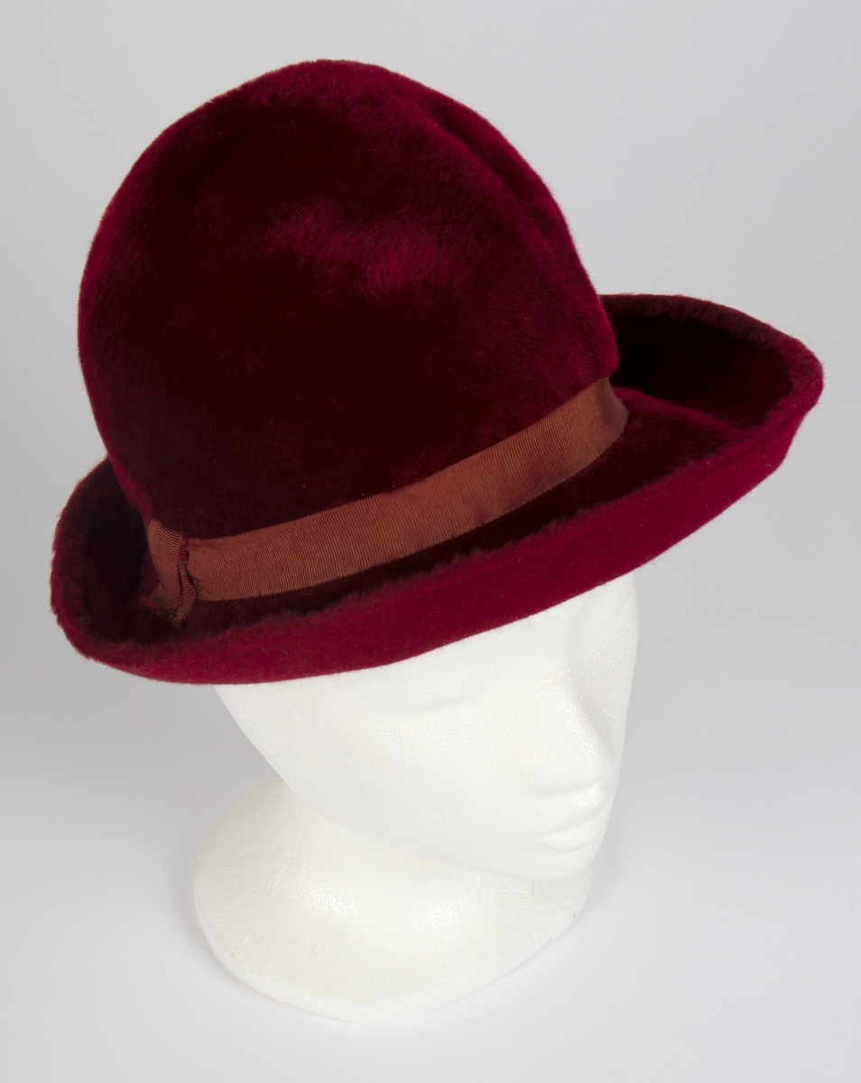 Håndsydd, burgunderrød fedora med høy, rund pull, nestet på innsiden, slik at den får to bulker på siden foran à la fedora herrehatter. Også toppen av den høye, runde pullen, er nestet på innsiden slik at den trekkes ned i en bulk. Relativt lang silkefloss. Smalt ripsbånd omkring pullen, med dekorativt element der båndet møtes bak på høyre side. Relativt bred brem, mykt oppoverbøyd og med smal falding på undersiden. Innvendig svettebånd av ripsbånd.