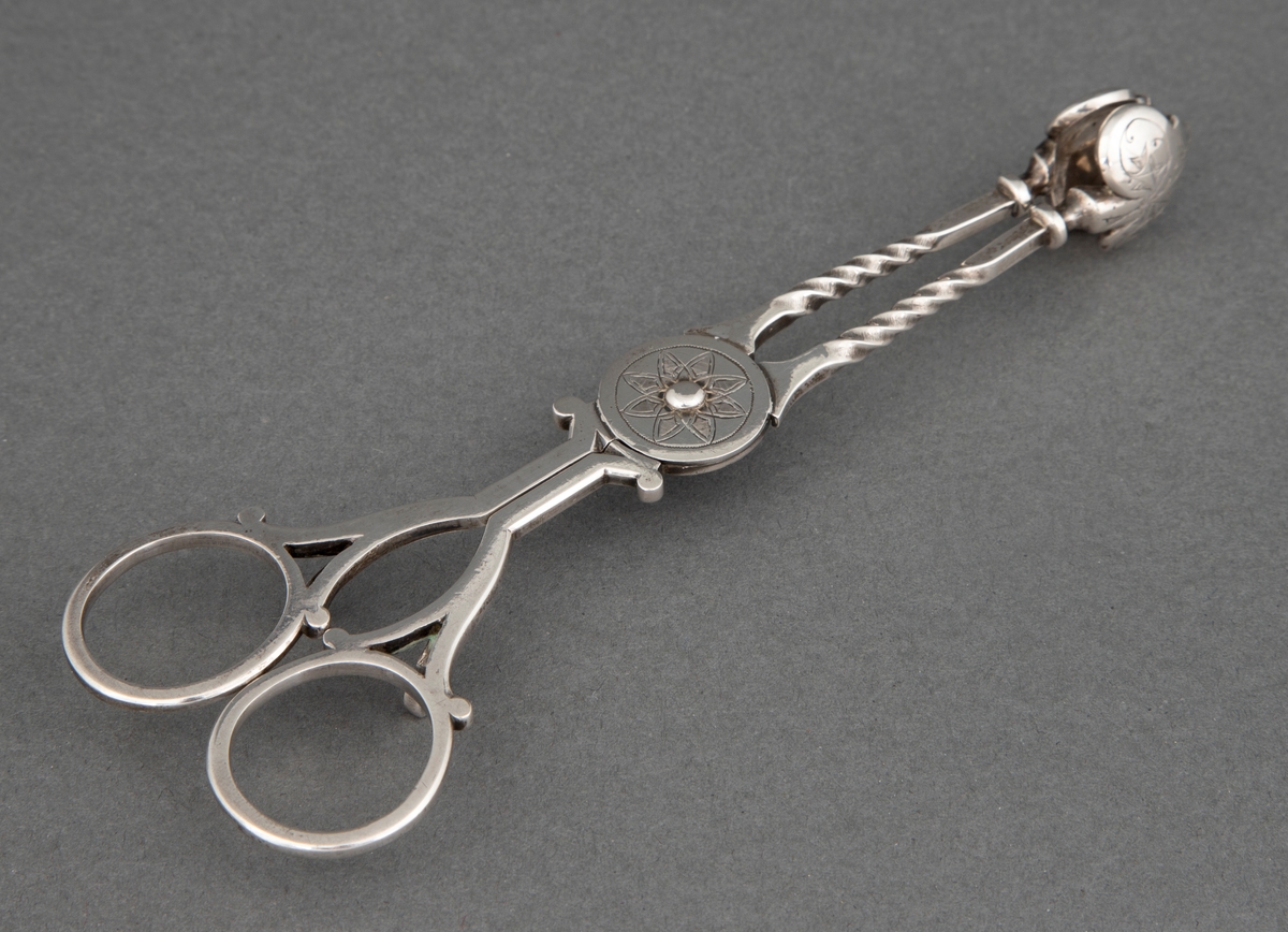 Sakseformet sukkerklype i sølv med sterkt profilerte, dreide tenger og løvformede skjeblad. Rund plate på aksen med blomstermotiv.