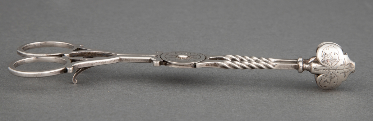 Sakseformet sukkerklype i sølv med sterkt profilerte, dreide tenger og løvformede skjeblad. Rund plate på aksen med blomstermotiv.