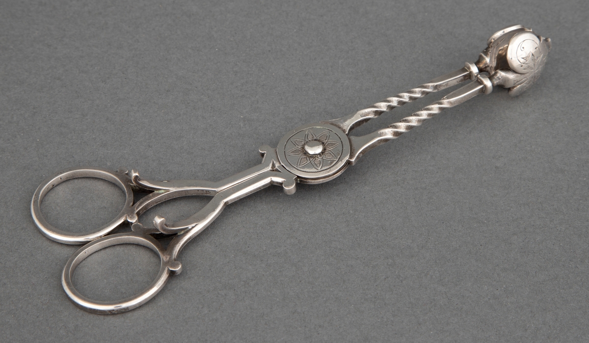 Sakseformet sukkerklype i sølv med sterkt profilerte, dreide tenger og løvformede skjeblad. Rund plate på aksen med blomstermotiv.