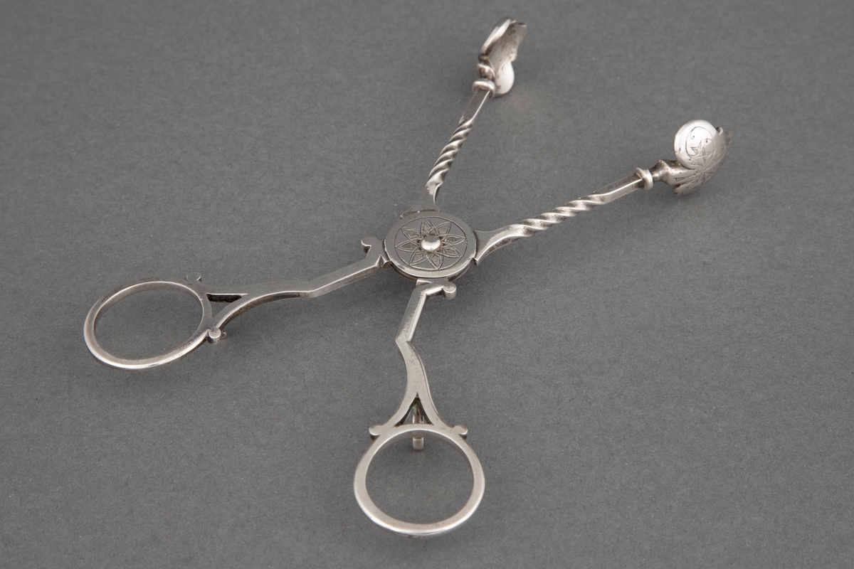 Sakseformet sukkerklype i sølv med sterkt profilerte, dreide tenger og løvformede skjeblad. Rund plate på aksen med blomstermotiv.