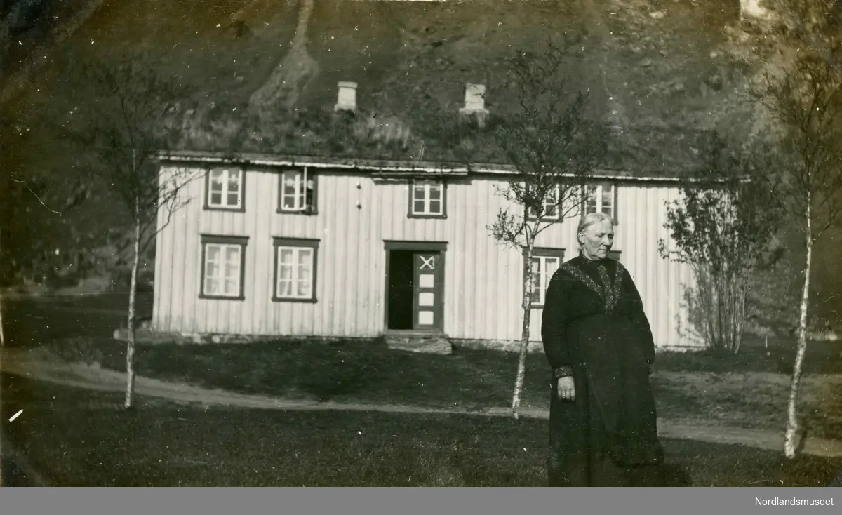 Eldre kvinne som står utenfor huset sitt. Kristianna Elisabet Karlsen (f. 1846). Kristianna var gift med Peder Markus Karlsen. Bak er våningshuset på Stegen gård.