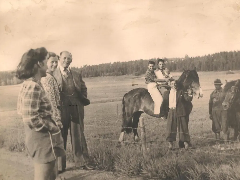 Sort-hvitt bilde av flere mennesker som står på en åker. På en hest til høyre sitter to personer, en gutt og en jente. Det er sommer.