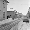 Parti av Follinegatan i Skänninge den snörika vintern 1959. Mellan adresserna 26 och senare rivna 24 tvekar ett par kampar inför gatans slaskföre. Redan där står ett modernare åk parkerat i form av en Ford Escort.