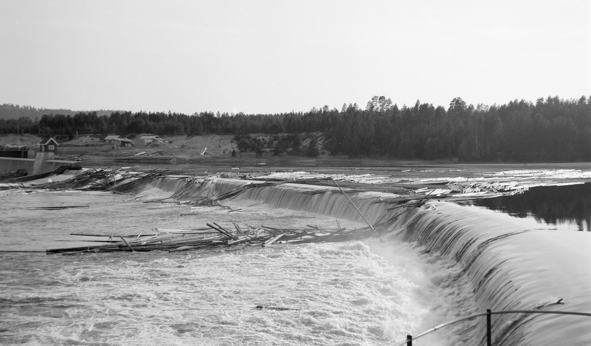 Kraftverksdammen ved Skjefstadfossen, lengst nord i Heradsbygda i Elverum. Fotografiet er tatt fra dammens landkar ved den østre elvebredden. Dammen var 312 meter lang, og den skulle stuve opp vann til kraftstasjonen, som lå litt lengre nede, ved vestre elvebredd (til venstre for venstre bildekant her). Fotografiet ble tatt i 1. mai 1932, på et tidspunkt da elva var flomstor. Dette innebar at det gikk vann over hele damkrona, ikke bare den østre enden, som var litt lavere enn den vestre fordi man ønsket at mest mulig av fløtingstømmeret skulle gå i den østre delen av elveløpet, som var djupest. Da dette fotografiet ble tatt hadde det samlet seg tømmerhauger, både på dammen og i fallsona nedenfor.

Skjefstadfossen hadde opprinnelig en fallhøyde på sju meter.  Ved bygginga av kraftverksdammen som dette fotografiet er tatt fra i 1909-1910 ble vannspeilet ovenfor Skjefstadfossen noe hevet, slik at fallhøyden ble økt til 10,5 meter.  Dette anlegget var det første store kraftverket som ble reist i den øvre delen av Glomma (nord for innsjøen Øyeren).