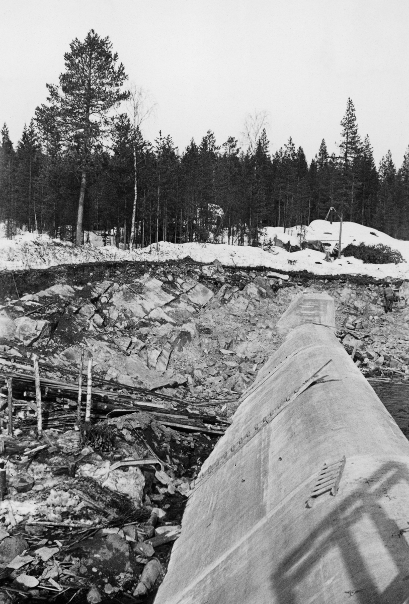 Bygging av dam for det nye kraftverket ved Kvernfallet i Søndre Osa, ei sideelv i Renavassdraget, som igjen er ei sidegrein i Glommavassdraget, i Åmot kommune i Hedmark. Fotografidt er tatt langsetter terskelen av armert betong, som skulle stuve opp vann til turbinrøret, som ble plassert langs søndre elvebredd. Dette fotografiet ble tatt mens anleggsarbeidet ennå pågikk, fra søndre mot nordre elvebredd. Forskalinga på muren var demontert, men det lå stiger på muren som gjorde det mulig for arbeiderne å klatre opp på murkrona. Anleggsarbeidet ble utført seint på vinteren og tidlig på våren, i en periode da vannføringa var minimal og kunne ledes til side for damkonstruksjonen.

Litt informasjon om kraftutbyggonga i Søndre Osa finnes under fanen «Opplysninger».