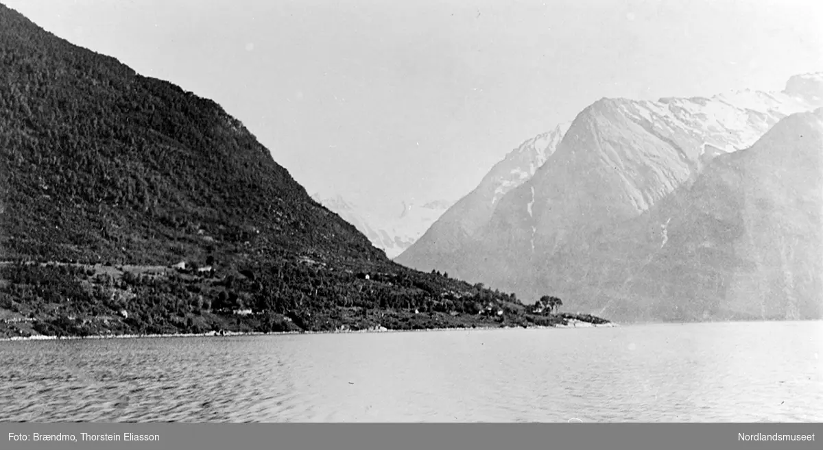 Stryn, Nordfjord, spredte bygninger på neset. Bratte fjell bak. I forgrunnen havet.