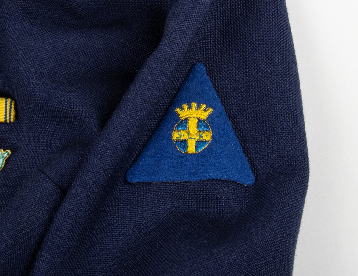 Uniform, klänning M/51 kv med tillhörande löstagbar krage, manschetter och skärp. Till klänningen medföljer en extra krage samt ett par manschetter. 
Monterade märken:
Riksförbundet Sveriges lottakårer S.L.K.
Släpspänne
Lottakårens orienteringsmärke i guld.
Lottakårens orienteringsmärke emaljerat.
Svenska lottakårens emblem S.L.K, broderat, fastsytt på vänster ärm.