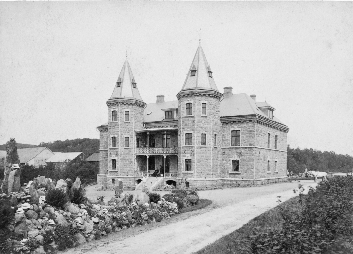 Från August Bondesons fotosamling. Mostorps slott i Getinge, uppfört 1878–80 efter ritningar av den danske arkitekten och formgivaren Thorvald Bindesböll (1846–1908). Byggherren Ludvig von Segebaden skänkte 1893 Mostorps Gård till sin yngsta dotter Ester och hennes man Rolf von Braun, med nyttjanderätt under hennes livstid. På verandorna sitter några personer, en med hund på övervåningen, och på trappan står nog godsherren själv i ljusa kläder och tropikhjälm. I förgrunden ett stenparti smyckat med stora snäckor och i bakgrunden till höger står ett par hästar förspända.