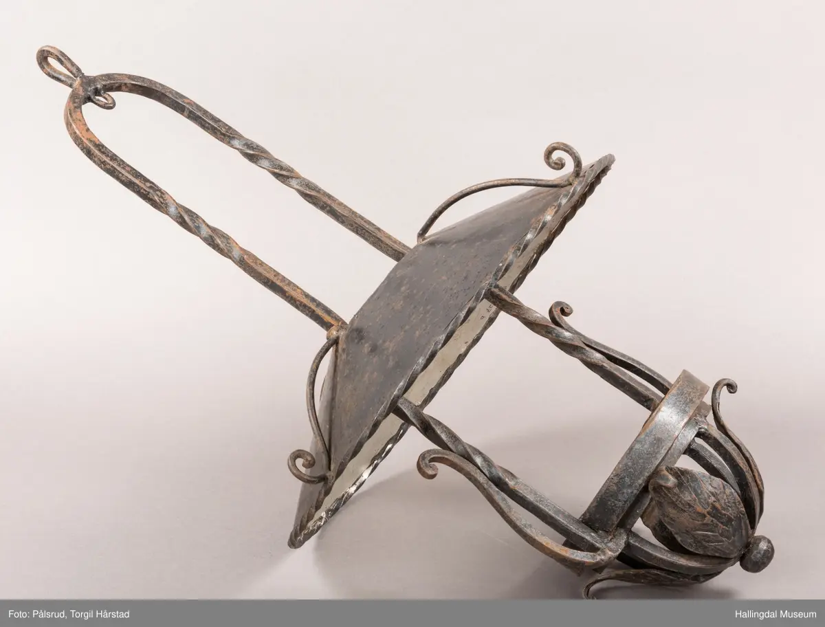 Lampeskjerm og holder til parafinlampe. Holderen er dekorert med fire jernblad. Skjermen er malt lys grå innvendig, sitter løst i opphenget. Opphenget ender i en bue hvor det er festet en ring til videre oppheng.