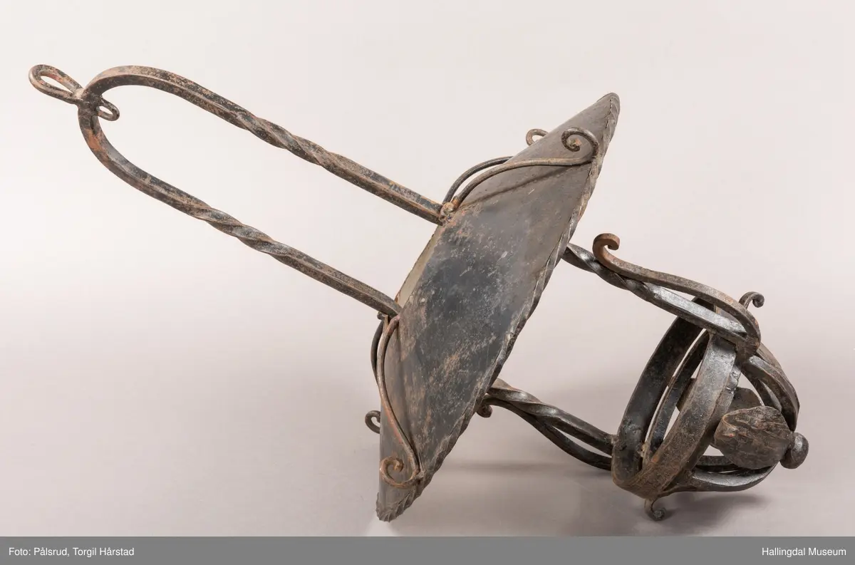 Lampeskjerm og holder til parafinlampe. Holderen er dekorert med fire jernblad. Skjermen er malt lys grå innvendig, sitter løst i opphenget. Opphenget ender i en bue hvor det er festet en ring til videre oppheng.