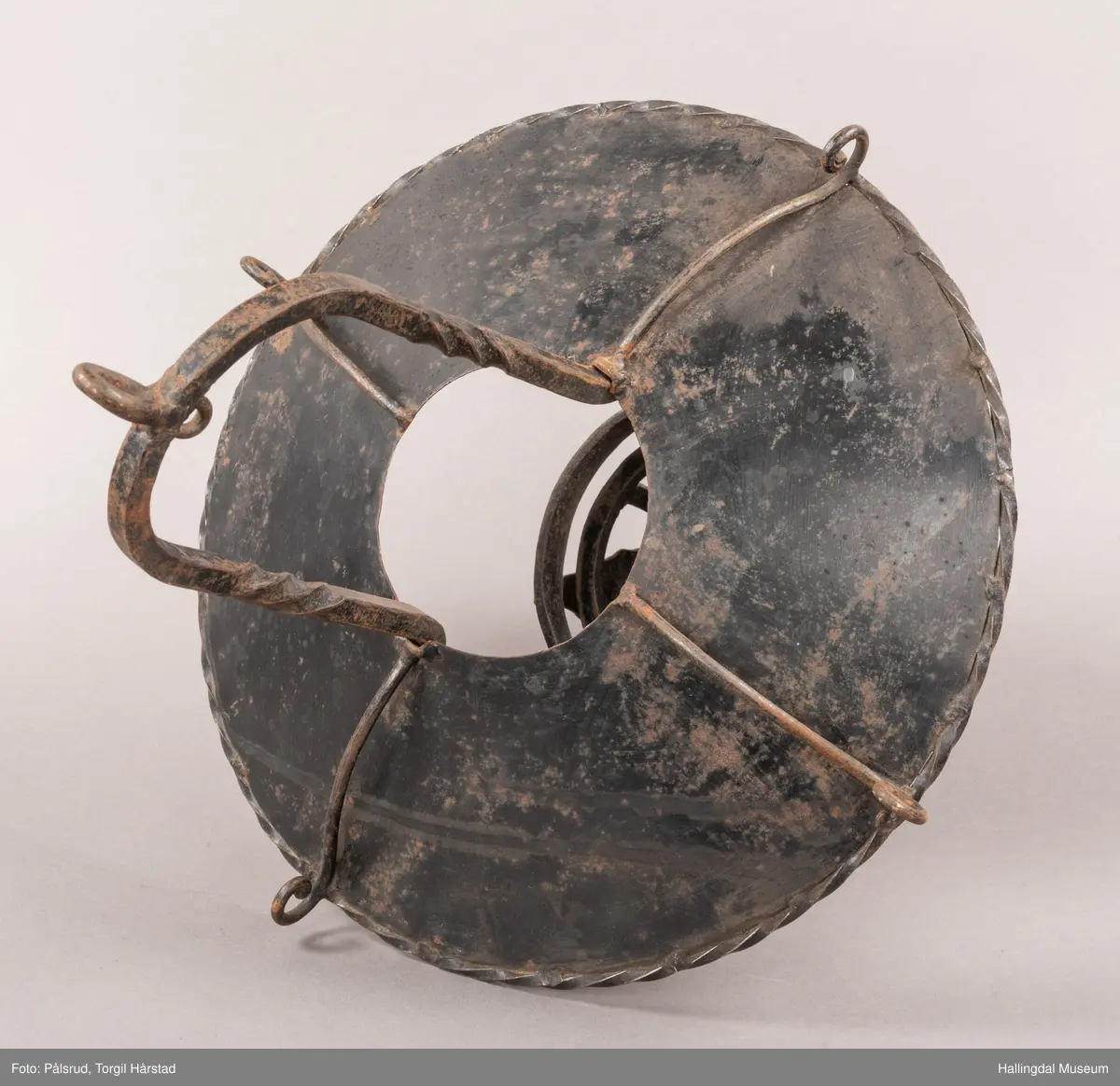 Lampeskjerm og holder til parafinlampe. Holderen er dekorert med fire jernblad. Skjermen er malt lys grå innvendig, sitter løst i opphenget. Opphenget ender i en bue hvor det er festet en ring til videre oppheng.