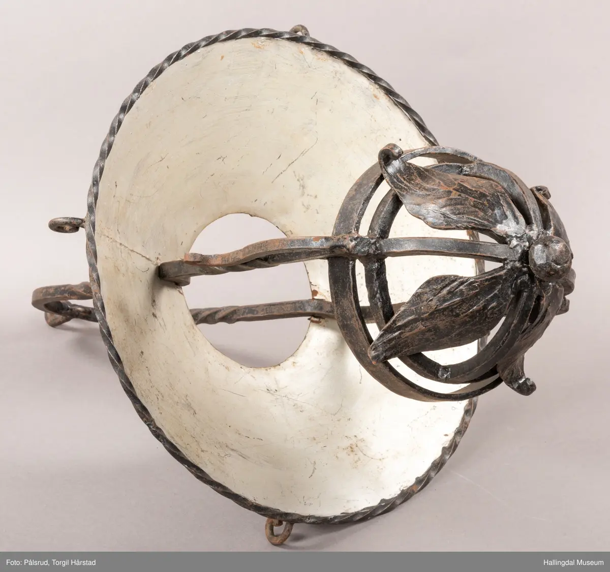 Lampeskjerm og holder til parafinlampe. Holderen er dekorert med fire jernblad. Skjermen er malt lys grå innvendig, sitter løst i opphenget. Opphenget ender i en bue hvor det er festet en ring til videre oppheng.