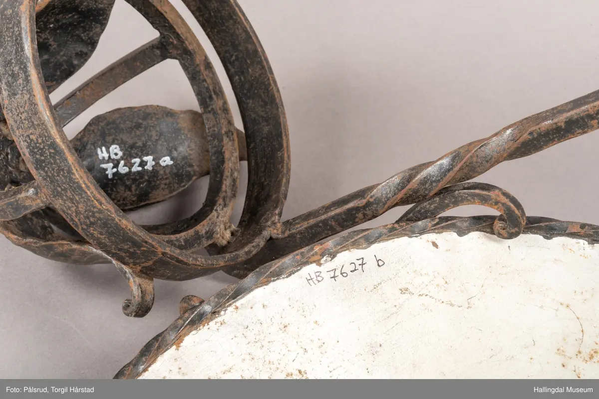 Lampeskjerm og holder til parafinlampe. Holderen er dekorert med fire jernblad. Skjermen er malt lys grå innvendig, sitter løst i opphenget. Opphenget ender i en bue hvor det er festet en ring til videre oppheng.