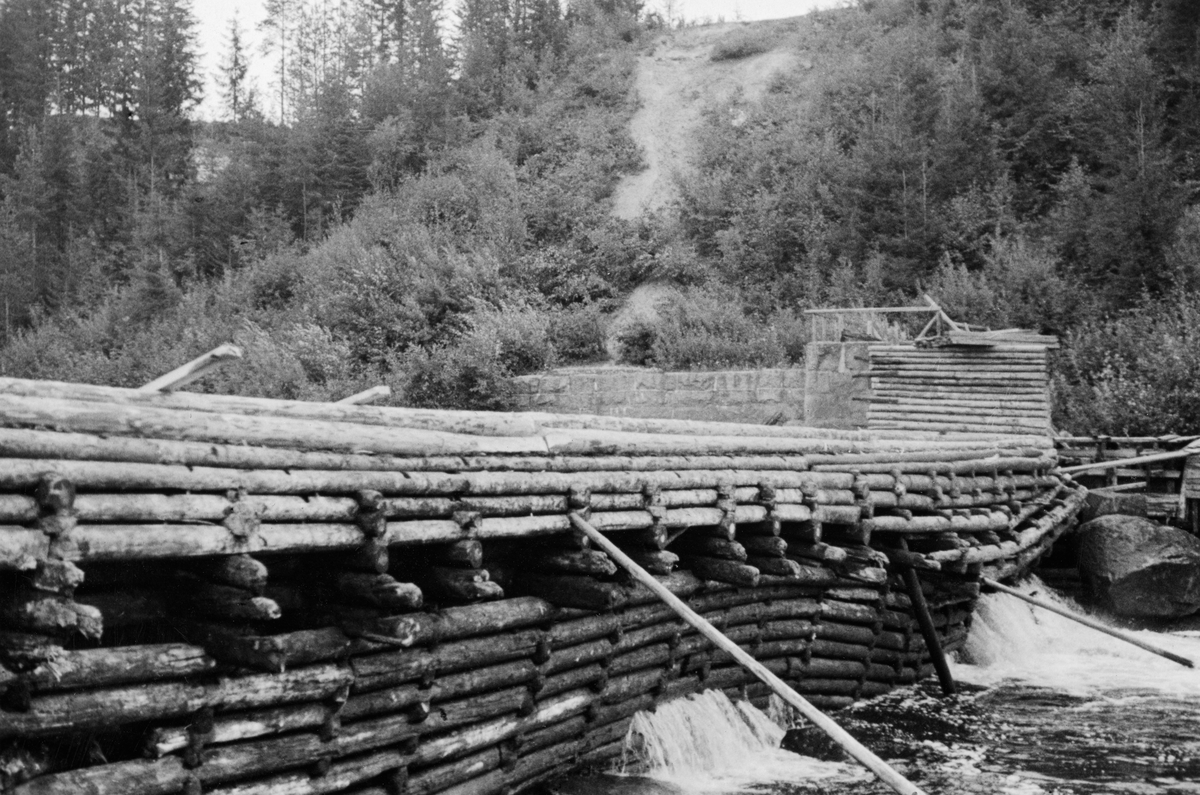 Syversætredammen, en tømmerkistedam i Flisa, ei sideelv til Glomma i Solør (Åsnes).  Fotografiet er tatt fra nordre elvebredd på skrå mot damkonstruksjonen.  Det viser en setning i konstruksjonen, antakelig ved djupålen i elva.  En del vann fosser gjennom tømmerkista, særlig der hvor setningen er kraftigst.