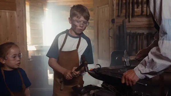 To barn iført forkle og beskyttelsesbriller deltar i smedarbeid i Snekkersmia på Norsk Folkemuseum. En gutt hamrer på et glødende jernstykke med en smed veiledende ved siden av, mens et yngre barn følger nøye med.