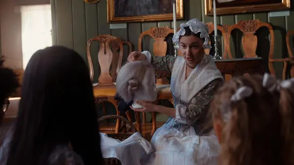 En dame i kostyme viser fram en parykk til to barn.