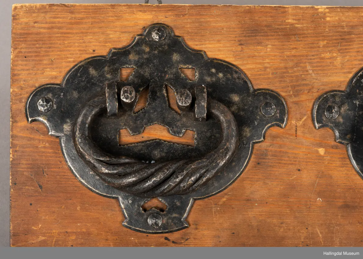 En treplate med to påmonterte dørbeslag, mulig dørhammere, i sort smijern. Det bakre beslaget er festet med metall-nagler, 4 stykk på hvert beslag. Selve håndtaket/hamrene har en oval form der endene er montert inn i to fester. En tykkere del av håndtakene er vridd. Platen har to kroker øverst til oppheng. Beslagene er festet med fire muttere på baksiden. 