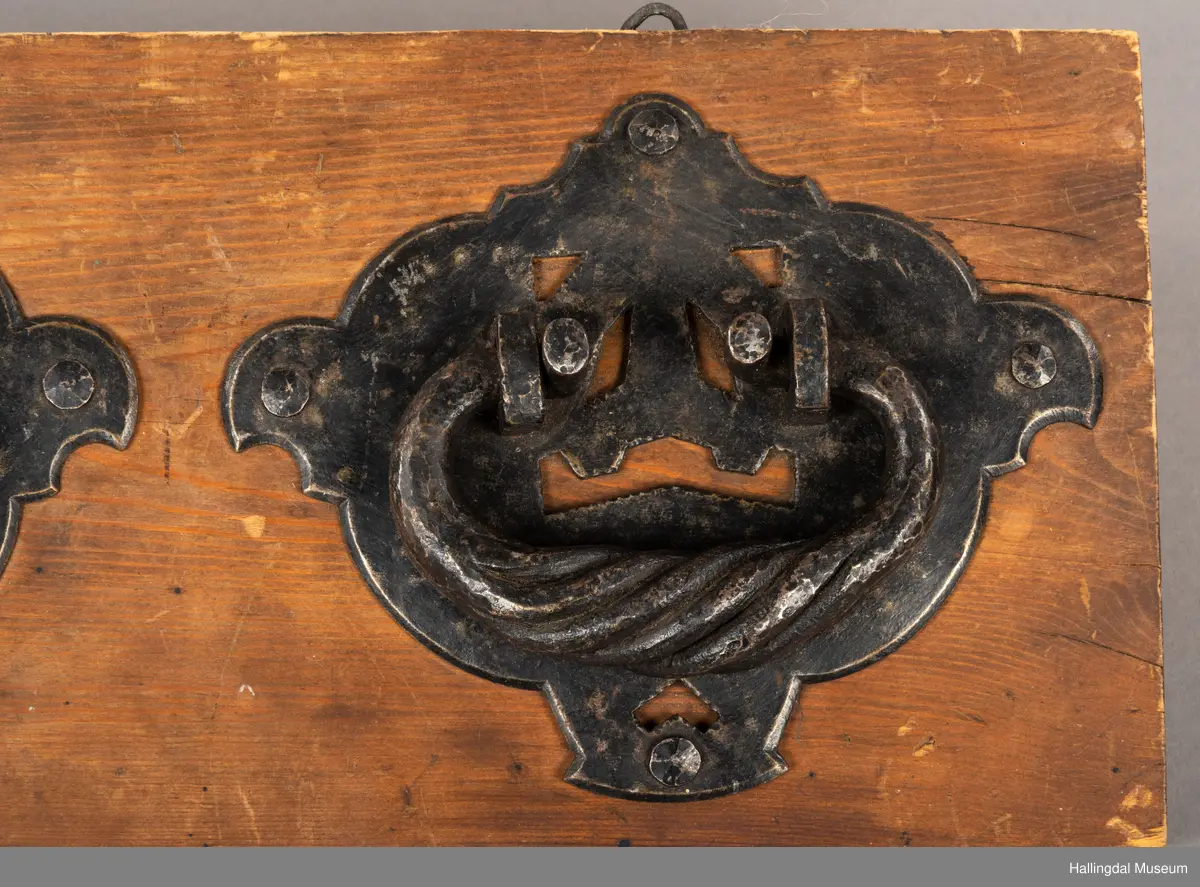 En treplate med to påmonterte dørbeslag, mulig dørhammere, i sort smijern. Det bakre beslaget er festet med metall-nagler, 4 stykk på hvert beslag. Selve håndtaket/hamrene har en oval form der endene er montert inn i to fester. En tykkere del av håndtakene er vridd. Platen har to kroker øverst til oppheng. Beslagene er festet med fire muttere på baksiden. 