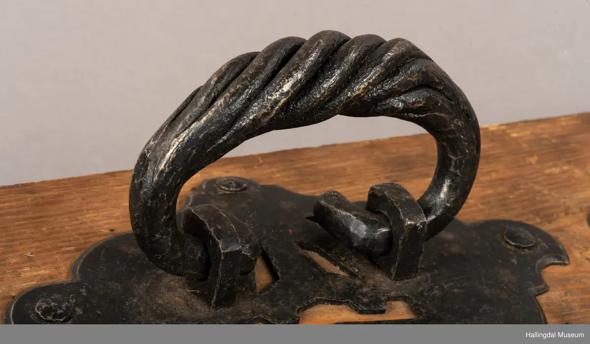 En treplate med to påmonterte dørbeslag, mulig dørhammere, i sort smijern. Det bakre beslaget er festet med metall-nagler, 4 stykk på hvert beslag. Selve håndtaket/hamrene har en oval form der endene er montert inn i to fester. En tykkere del av håndtakene er vridd. Platen har to kroker øverst til oppheng. Beslagene er festet med fire muttere på baksiden. 