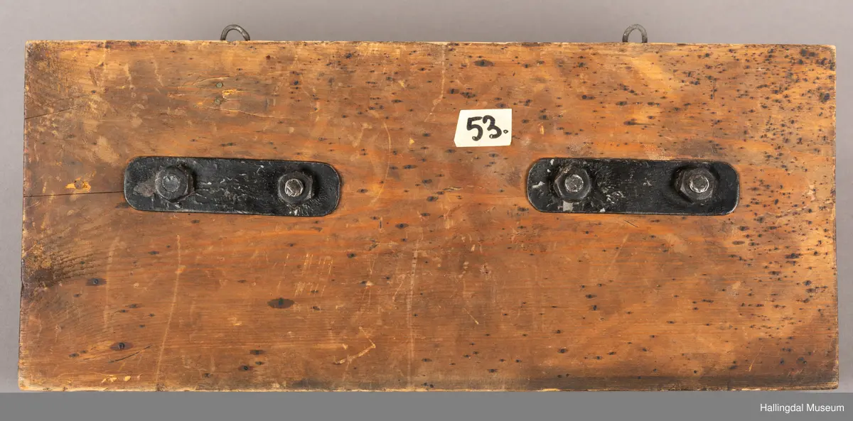 En treplate med to påmonterte dørbeslag, mulig dørhammere, i sort smijern. Det bakre beslaget er festet med metall-nagler, 4 stykk på hvert beslag. Selve håndtaket/hamrene har en oval form der endene er montert inn i to fester. En tykkere del av håndtakene er vridd. Platen har to kroker øverst til oppheng. Beslagene er festet med fire muttere på baksiden. 