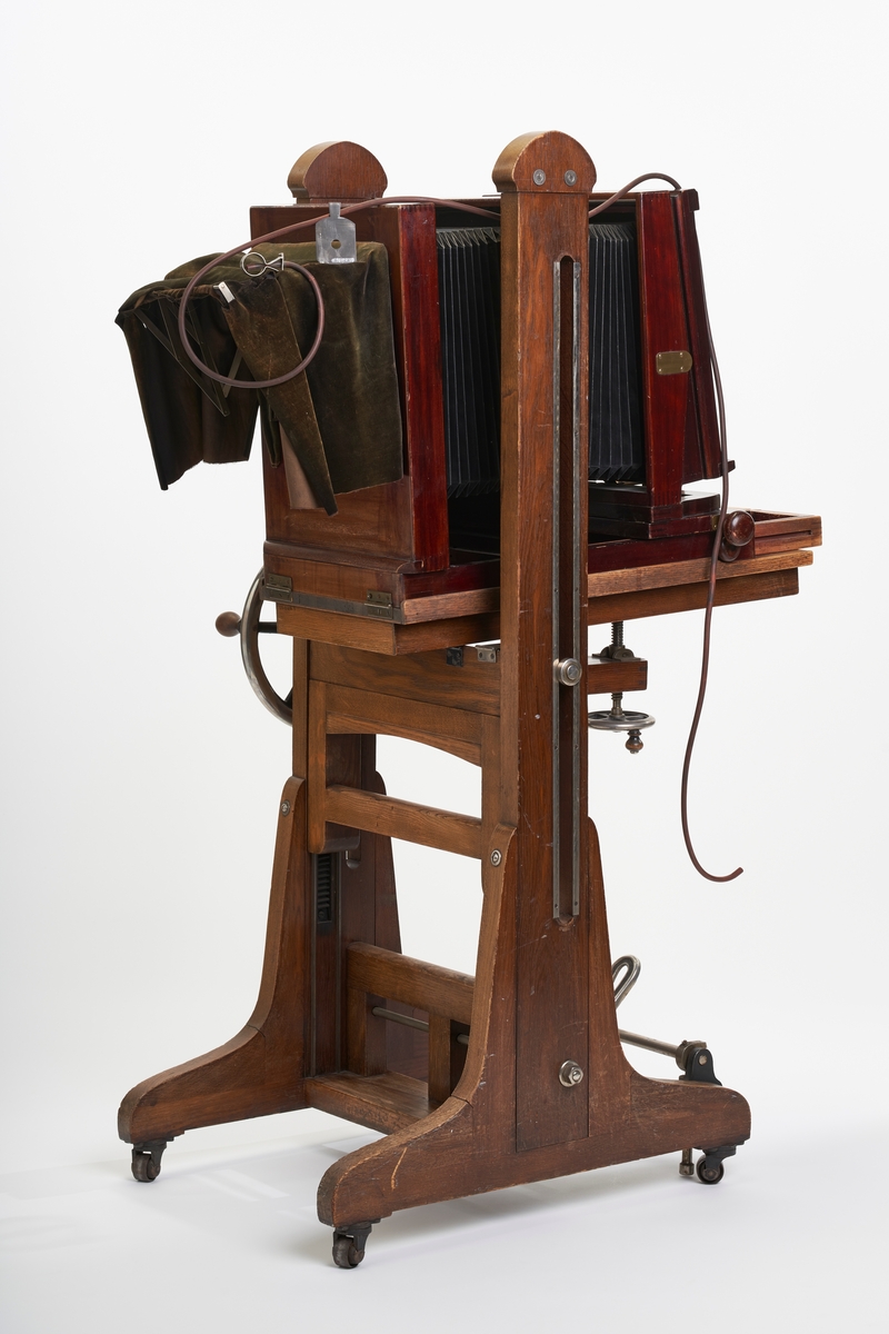 Fotografiapparat på stativ. Stativet er massivt og har justerbar sokkel. Både fotografiapparatet og stativet er i tre. Stativet står på hjul. Fotografiapparatet har uttrekkbar belg, men mangler frontplate og opptikk. Det hører med en slange til fotografiapparatet og en innstikksmodul for å velge blenderåpnimg på motivet.
